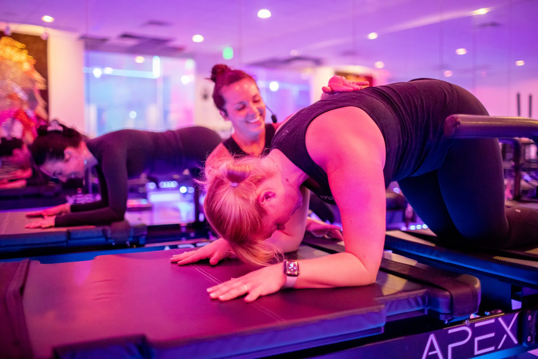 A smiling APEX teacher with a hand on a student's back as she stretches forward on the Xformer under pink studio light